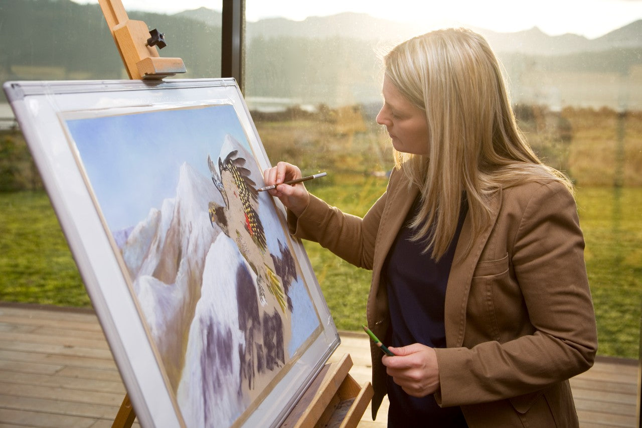 Karen painting an animal on paper set on an easel, with a scenic outdoor landscape visible through the window behind her.