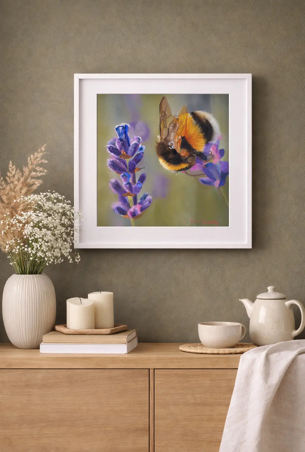 Framed artwork of a bee on lavender hanging above a wooden cabinet with decorative items.
