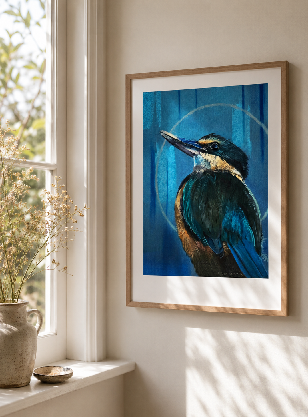 Kotare Sacred Kingfisher print in a raw wood frame on a wooden table, styled with a paintbrush, ceramic mug and linen cloth in a warm studio setting
